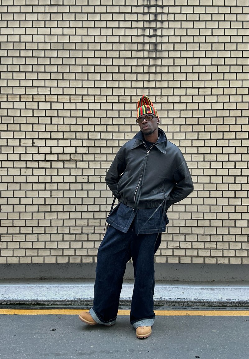 Veste en cuir noir, pantalons en denim surdimensionnés, sabots beige et un chapeau rayé coloré. Adossé à un mur de briques claires.