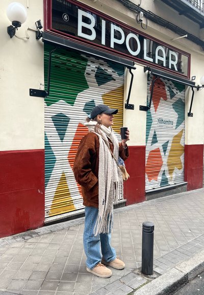 Chaqueta oversize de ante marrón, bufanda gris claro, vaqueros azules, zapatos slip-on en color beige. Fondo de tienda geométrico y colorido con letrero "BIPOLAR".