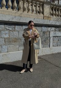 Manteau beige avec un grand col, assorti à un pantalon noir et des ballerines crème. Un sac blanc est porté sur l'épaule, contre un fond en pierre.
