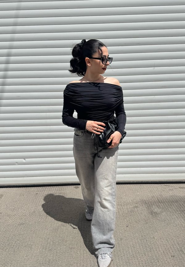 Top preto, com ombros à mostra, franzido, combinado com jeans de perna larga em cinza claro. Acessórios incluem uma mala preta e anéis grossos. Luz do sol projetando sombras.