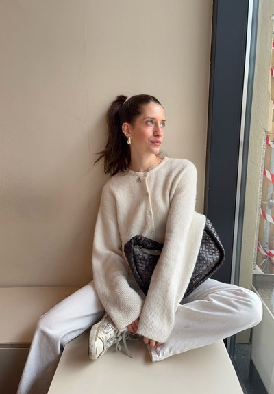 Joven mujer con coleta oscura, vestida con suéter crema y pantalones blancos, sentada con las piernas cruzadas junto a la ventana sosteniendo un bolso negro tejido.