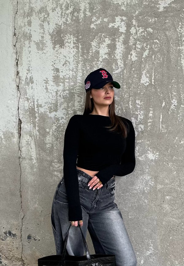 Schwarzes Langarmoberteil und ausgebleichte schwarze Jeans, kombiniert mit einer marineblauen Kappe mit rotem Logo. Eine schwarze Tragetasche wird in einer Hand gehalten.