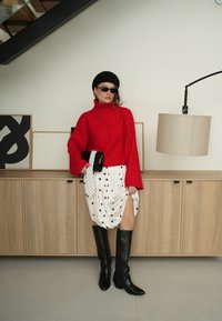 Femme portant un pull à col roulé rouge, une jupe blanche à pois avec une fente, des bottes noires montantes jusqu'aux genoux, un béret noir et des lunettes de soleil, debout à l'intérieur.