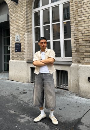 Camisa de manga corta beige claro sobre una camiseta blanca, pantalones cortos grises de pierna ancha, calcetines blancos y zapatos beige, con un fondo de pared de ladrillo.