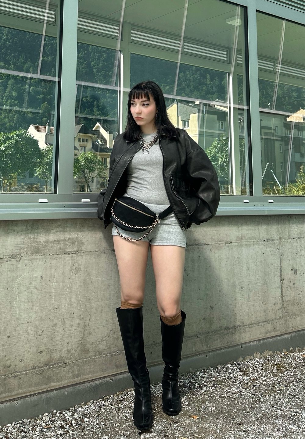 Schwarze Lederjacke, graues enges Oberteil, graue Shorts und kniehohe schwarze Stiefel. Schwarze Hüfttasche mit Kettenakzent. Urbane Umgebung mit Grünpflanzen.