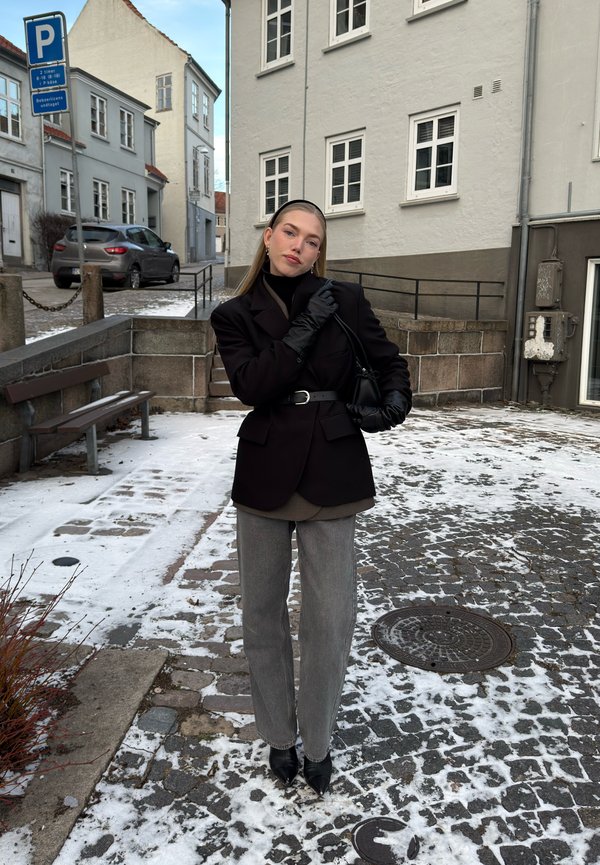 Mlada ženska, oblečena v črni blazer, rokavice in trak za glavo, stoji na zasneženi tlakovani ulici v evropskem mestu.