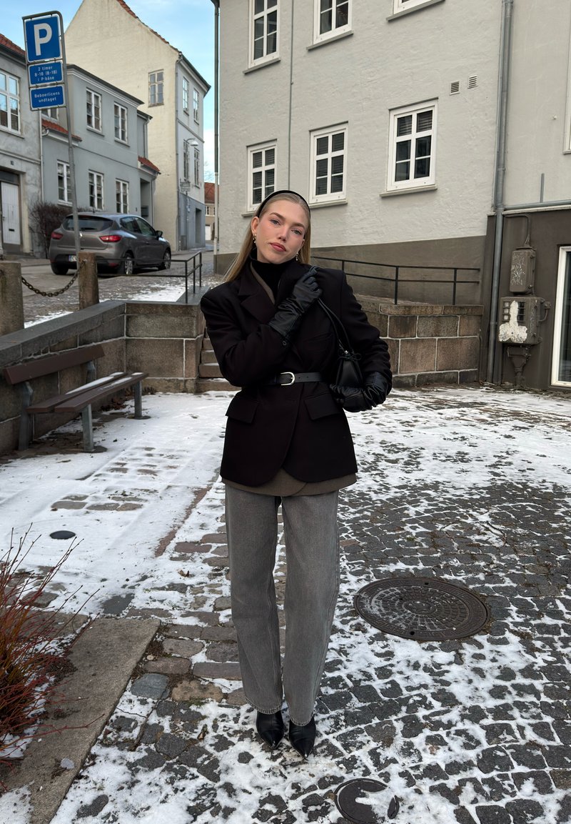 Jeune femme portant un blazer noir, des gants et un bandeau, debout dans une rue pavée enneigée d'une ville européenne.