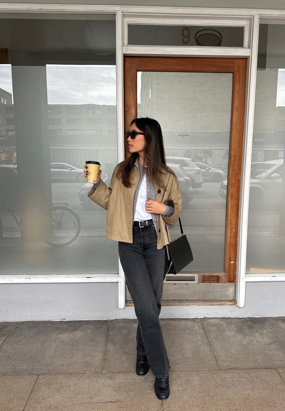 Beige cropped jakke over grå skjorte, sorte højtafsnittede jeans, sorte støvler, holder en kaffekop, står ved en glasskydedør.