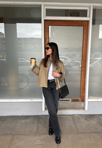 Beige cropped jakke over grå skjorte, sorte højtafsnittede jeans, sorte støvler, holder en kaffekop, står ved en glasskydedør.