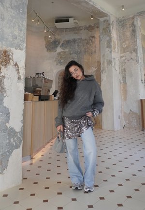 Mujer con cabello largo, sudadera con capucha gris, falda con lentejuelas, jeans anchos de tono claro, zapatillas deportivas y bolso gris, de pie en el interior de un café con paredes de textura desgastada.