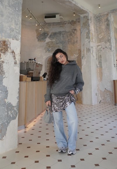 Mujer con cabello largo, sudadera con capucha gris, falda con lentejuelas, jeans anchos de tono claro, zapatillas deportivas y bolso gris, de pie en el interior de un café con paredes de textura desgastada.