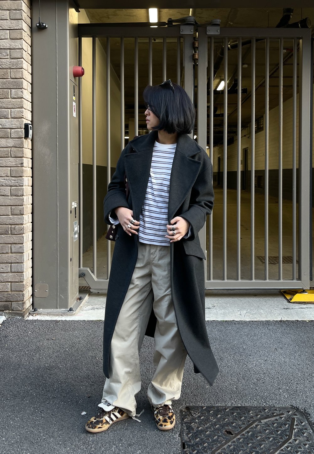 Personne aux cheveux courts portant un long manteau sombre, une chemise rayée, un pantalon beige et des chaussures à imprimé léopard, debout devant une entrée à grille métallique.