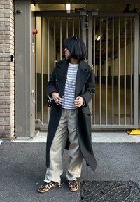 Personne aux cheveux courts portant un long manteau sombre, une chemise rayée, un pantalon beige et des chaussures à imprimé léopard, debout devant une entrée à grille métallique.