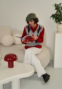 Ένα άτομο διαβάζει ένα περιοδικό σε έναν καμπυλωτό μπεζ καναπέ, φορώντας μια κόκκινη μπλούζα με γκρι αργυρό γιλέκο, λευκά παντελόνια και μαύρες μπότες.