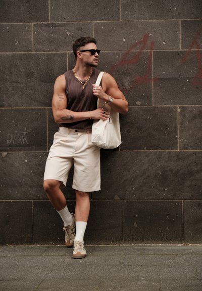 Pantalones cortos beige, top sin mangas marrón, calcetines blancos y zapatillas beige. Los accesorios incluyen collares y un bolso tipo tote. De pie contra una pared de piedra.