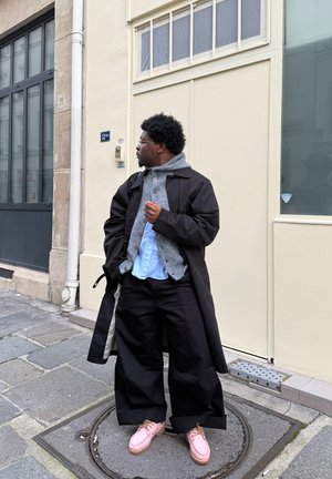 Abrigo largo negro, suéter texturizado gris claro, camisa azul y pantalones sueltos negros. Zapatos rosas y de pie contra una pared de color claro.