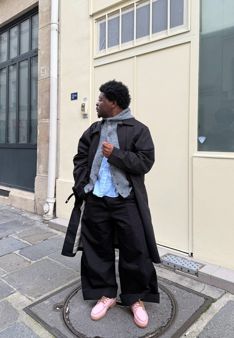 Manteau long noir, pull texturé gris clair, chemise bleue et pantalon noir ample. Chaussures roses, debout contre un mur de couleur claire.