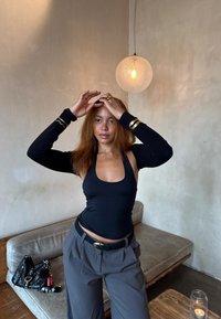 Mulher em top halter preto e calças cinzentas arruma o cabelo numa sala minimalista com uma luz pendente, sofá, bolsa, óculos de sol e vela na mesa.