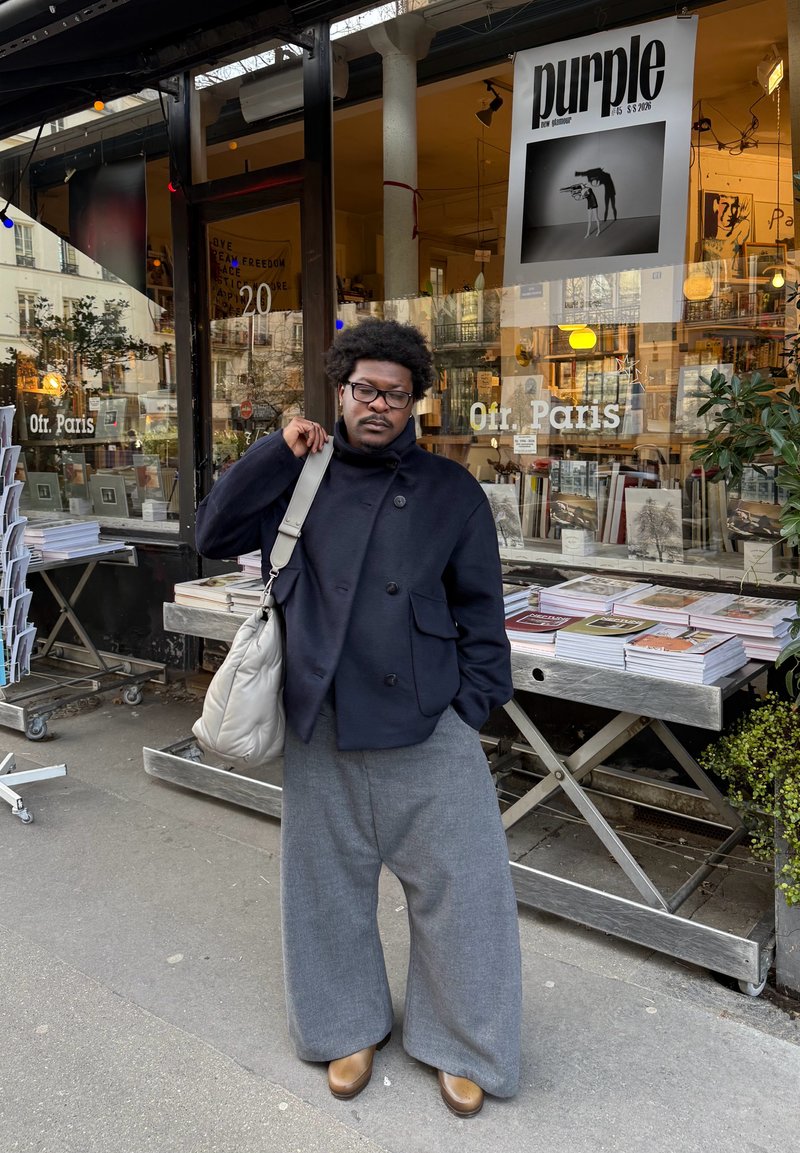 Άνδρας με γυαλιά και σγουρά μαλλιά φοράει μαύρο σακάκι και φαρδιά γκρι παντελόνια, κρατώντας μπεζ τσάντα, στέκεται έξω από βιβλιοπωλείο με βιβλία στην έκθεση.