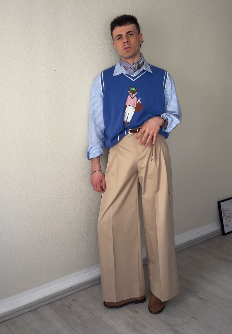Jeune homme portant un gilet bleu brodé de renard sur une chemise rayée, pantalon beige à jambes larges, debout à l'intérieur contre un mur uni.