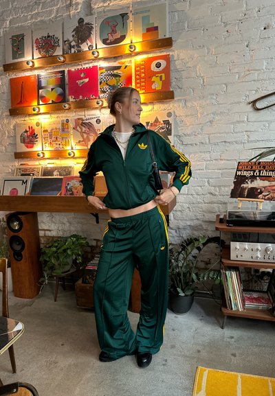 Chándal atlético verde con rayas amarillas, que incluye una chaqueta con cremallera y pantalones de cintura elástica. Unas zapatillas negras y un bolso pequeño completan el look.