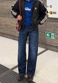 Veste noire zippée avec des rayures blanches, sweat à capuche bleu, jean bleu à jambes larges et chaussures marron. Petit sac bandoulière bordeaux.