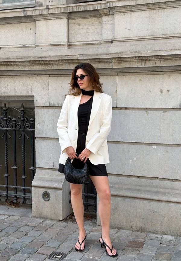 Donna che indossa un blazer bianco, un vestito nero, occhiali da sole neri e una borsa nera, in piedi contro un muro di pietra con una recinzione in ferro nelle vicinanze.