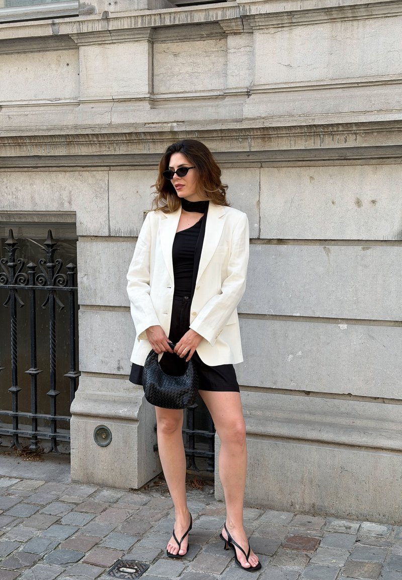 Frau trägt weißen Blazer, schwarzes Kleid, schwarze Sonnenbrille und schwarze Handtasche und steht an einer Steinmauer mit einem nahegelegenen Eisenzaun.
