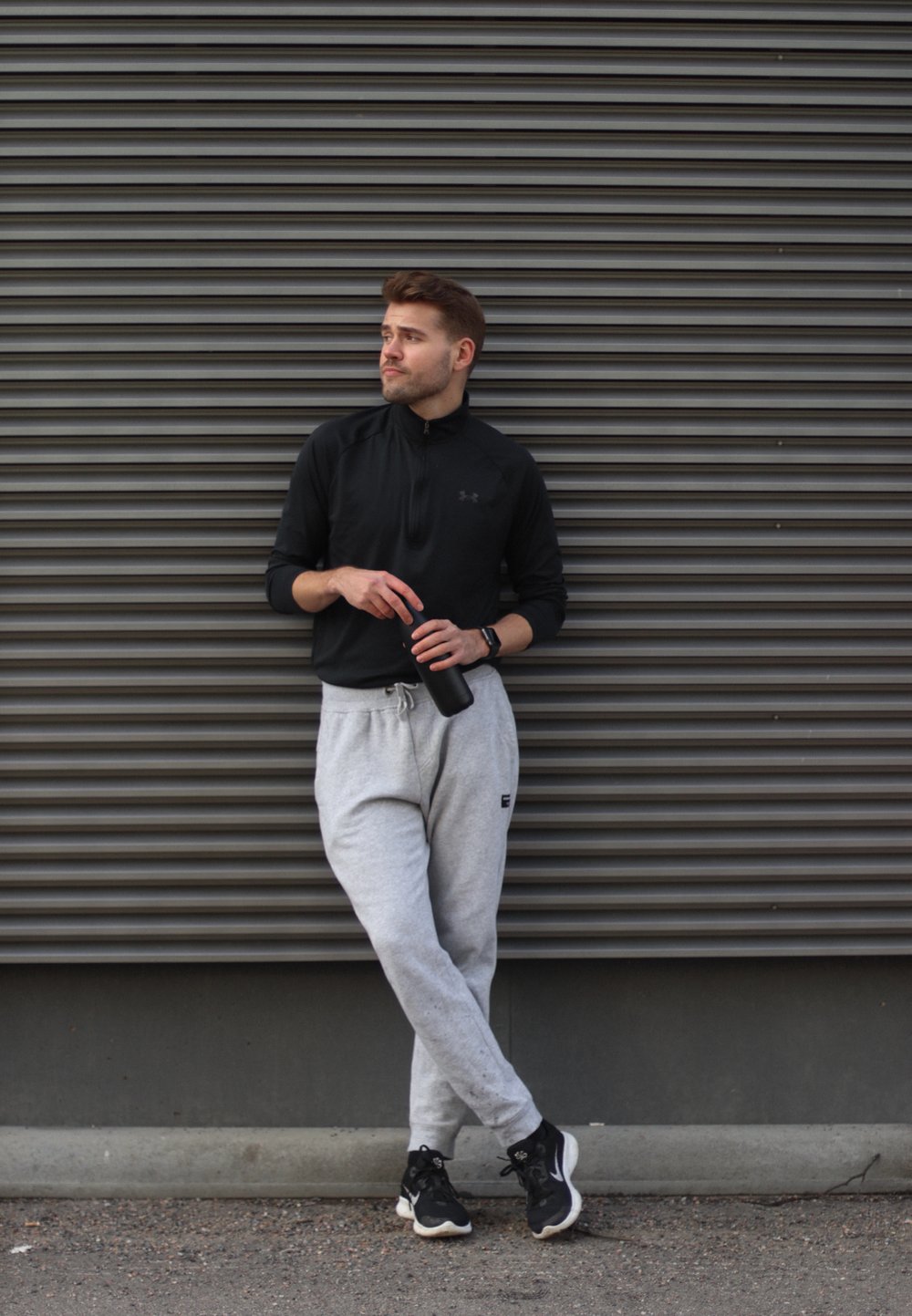 Maglietta sportiva nera con zip, pantaloni della tuta grigi con logo, bottiglia d'acqua nera, scarpe da ginnastica nere. In piedi contro un muro grigio con texture.