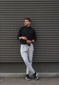 Maglietta sportiva nera con zip, pantaloni della tuta grigi con logo, bottiglia d'acqua nera, scarpe da ginnastica nere. In piedi contro un muro grigio con texture.