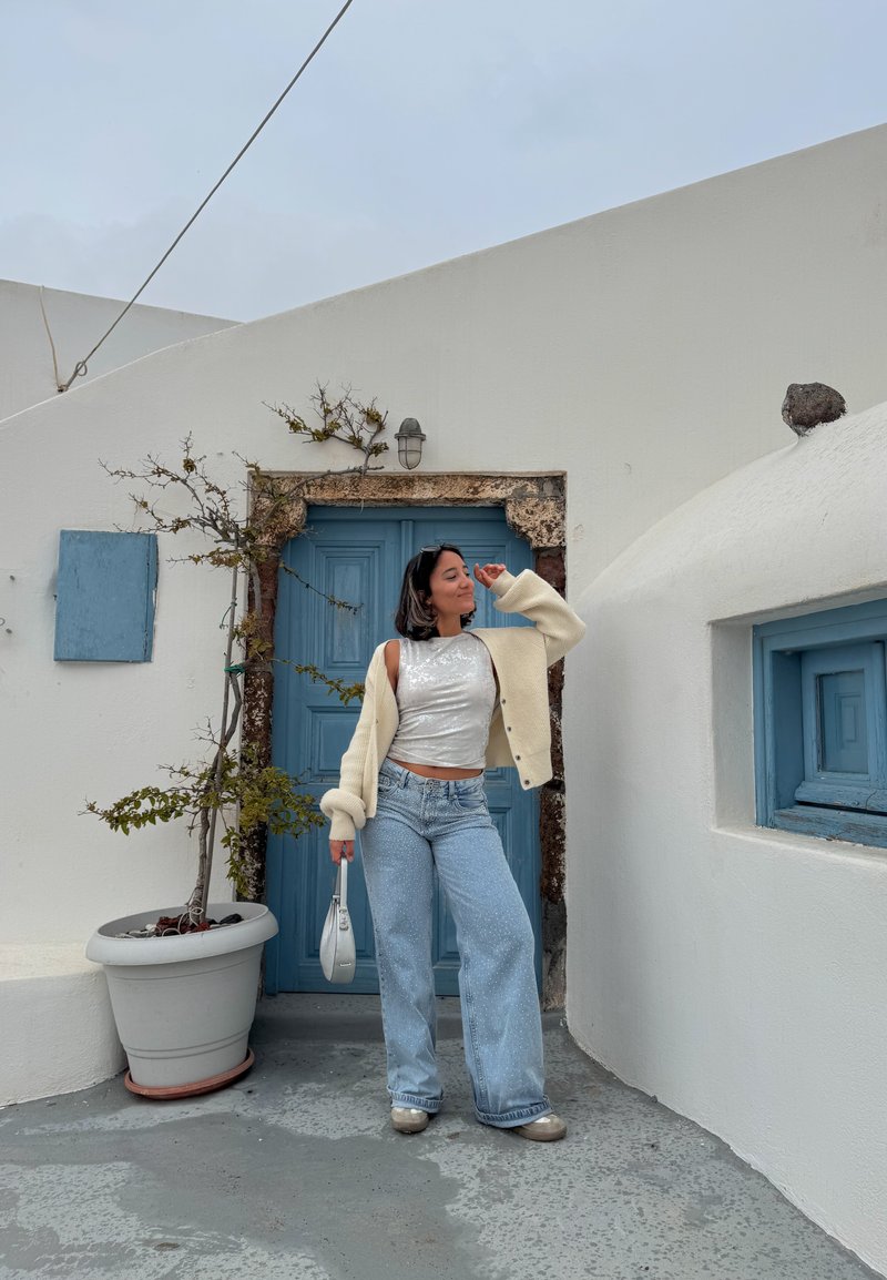 Femme en haut argenté, cardigan crème et jean large posant près d'une porte bleue avec une plante en pot devant un bâtiment blanc sous un ciel gris.