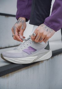 Ljusgrå löparsko med lila "N"-logotyp, texturerad meshovandel och klumpig vit sula. Snörena justeras på en trappa.