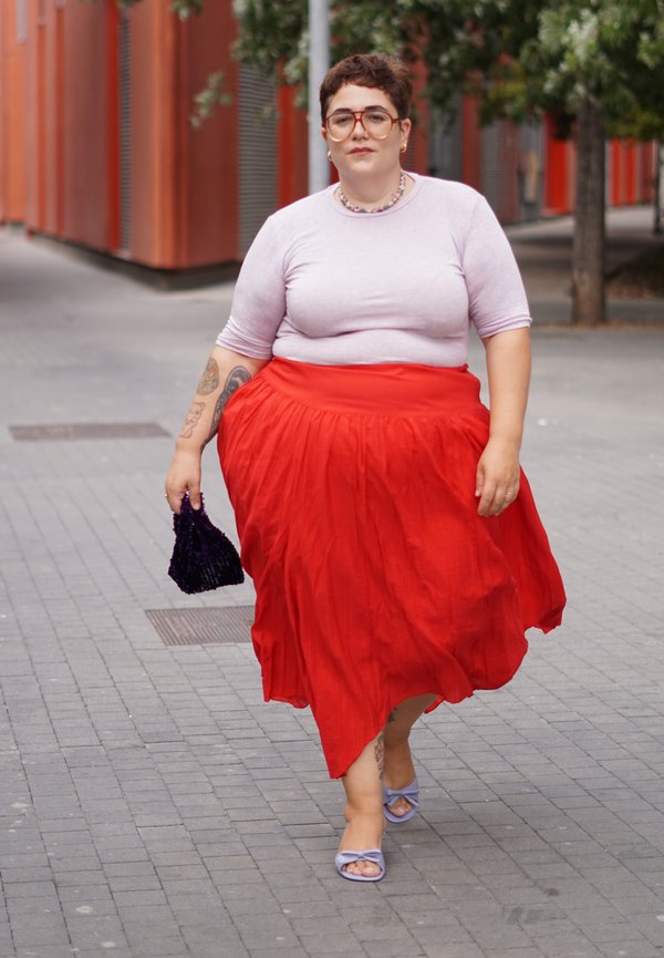 Person med kort hår iført briller, en lyserød top, rød nederdel og lilla sandaler, går på en byfortov og holder en lille sort taske.