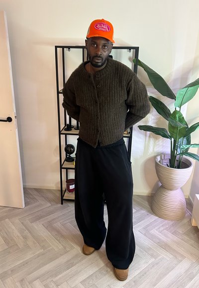 Cárdigan marrón acanalado con botones, pantalones amplios negros y calzado marrón claro. Gorra naranja con logo. Entorno interior con planta verde y estantería.