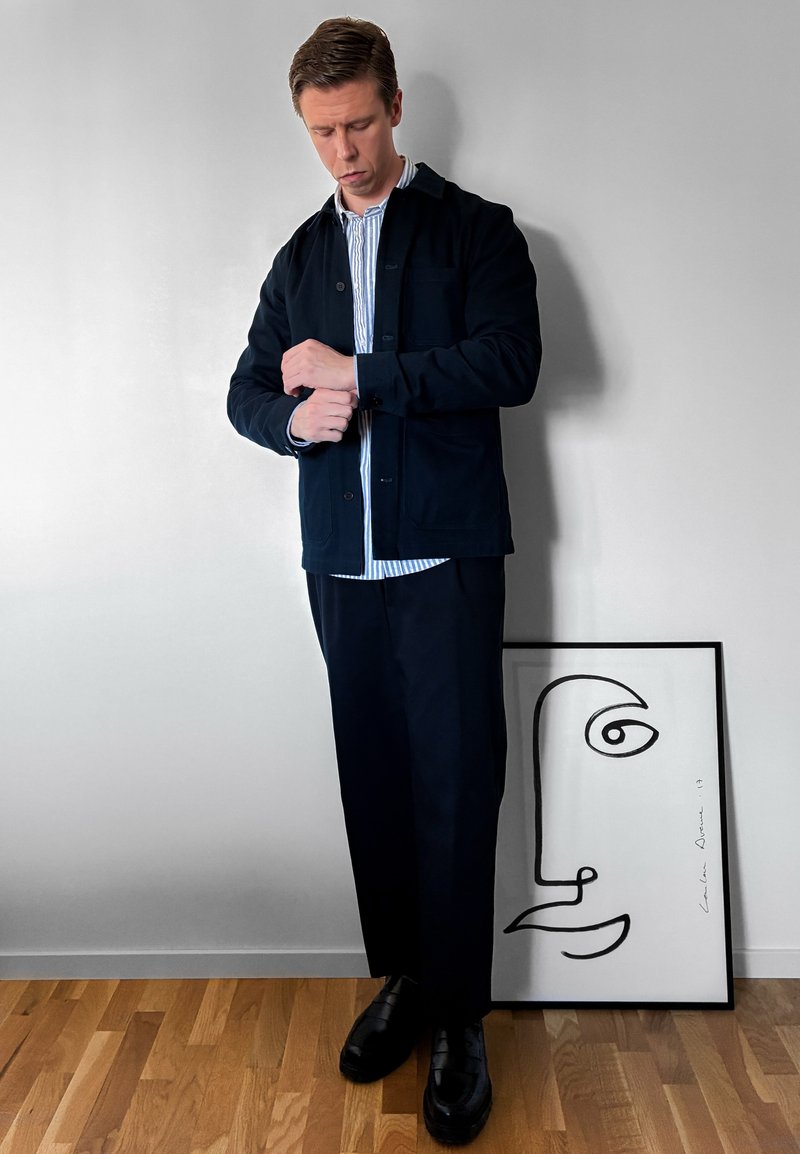Homme en veste et pantalon foncés ajustant la manche, debout à côté d’un visage en art linéaire minimaliste encadré sur un sol en bois contre un mur blanc.