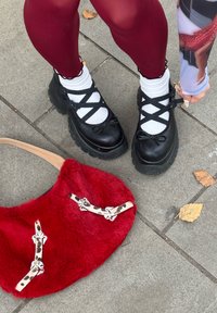 Čierne platformové topánky s prekríženými remienkami cez biele ponožky; červená chlpatá kabelka s dekoratívnymi remienkami na chodníku.
