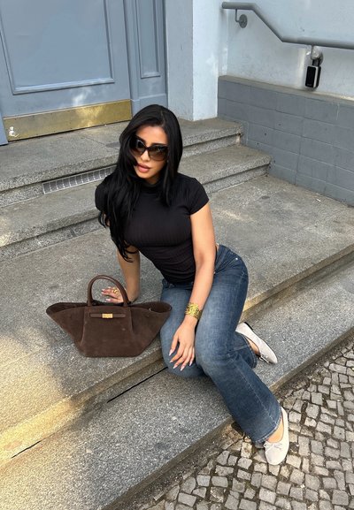 Mujer con cabello largo y oscuro, gafas de sol, top negro y vaqueros sentada en unas escaleras de piedra junto a un gran bolso marrón en un entorno urbano.