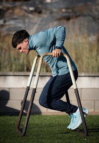 Personne effectuant des dips sur des barres métalliques, portant un haut à fermeture éclair bleu clair et un pantalon bleu marine, avec de l'herbe texturée et un mur en pierre en arrière-plan.