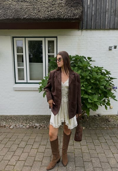 Mujer con blazer de ante marrón, vestido de encaje blanco, botas marrón hasta la rodilla y gafas de sol, se encuentra en un camino de adoquines junto a una casa de ladrillo blanco y un arbusto verde.