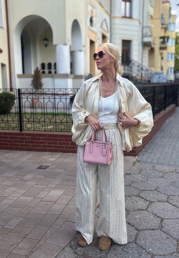 Kobieta ubrana w oversize'ową kremową bluzkę, szerokie spodnie w paski, okulary przeciwsłoneczne, trzymająca małą różową torebkę, stoi na chodniku w pobliżu budynków.