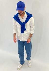Biela košeľa na gombíky, modrý sveter zaviazaný okolo ramien, svetlomodré džínsy a biele tenisky. Modrá šiltovka s detailom loga.