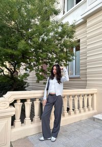 Ανοιχτό γκρι ζακέτα, λευκό μπλουζάκι, σκούρο γκρι παντελόνι με φαρδιά πόδια και λευκά αθλητικά παπούτσια με μαύρες λωρίδες. Στηρίζεται σε μια διακοσμητική κουπαστή, με φόντο πράσινο δέντρο.