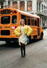 Kollane puffer-jope, põlveni triibuline kleit, mustad sukkpüksid ja pahkluu saapad. Sinine käekott. Taustal kollane koolibuss.