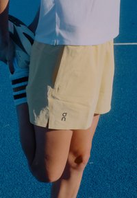 Gaiši dzelteni sporta šorti ar sānu spraugu, apakšējā kreisajā pusē ar logotipu. Izstrādāti ērtībai un kustībai sporta aktivitāšu laikā.