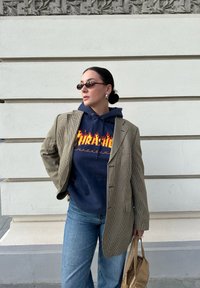 Καφέ σχέδιο καρό σακάκι πάνω από μπλε φούτερ με φλόγες, μπλε τζιν και καφέ τσάντα ώμου. Στρογγυλά γυαλιά ηλίου και εντυπωσιακά σκουλαρίκια.