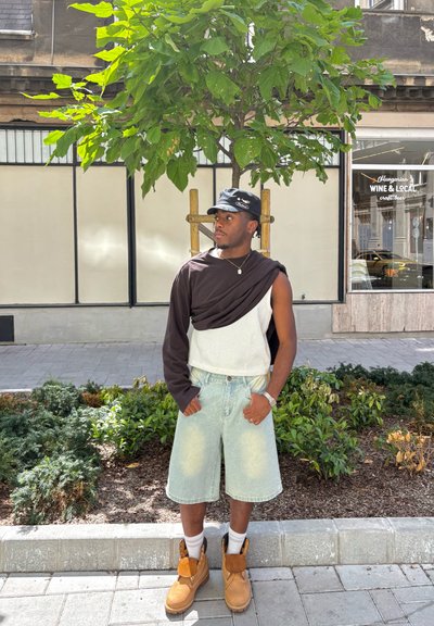 Una persona está de pie al aire libre vistiendo una camiseta negra de manga larga sobre una blusa blanca de punto, pantalones cortos de mezclilla claros y botas marrones con calcetas blancas.