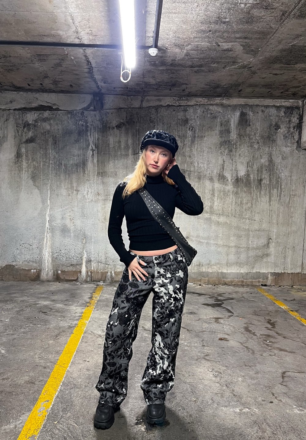 Black textured sweater, patterned black and white wide-leg pants, black cap, and a studded black crossbody bag in a concrete garage setting.