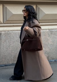 Haină maro, bluză neagră și pantaloni. Țin în mână o geantă de mână din piele intoarsă maro închis, cu mâner scurt și detaliu argintiu. Pe trotuar.