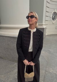 Schwarze, kurze Jacke mit verzierten Knöpfen, cremefarbene Bluse, schwarzer Rock und eine goldene, mit Perlen verzierte Handtasche mit Fransen. Schwarze Sonnenbrille komplettiert den Look.
