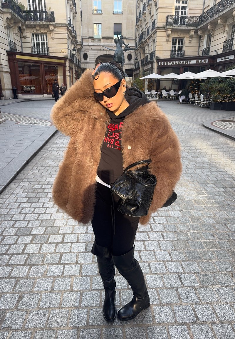 Femme portant un grand manteau en fourrure marron, des lunettes de soleil noires, un sweat à capuche noir, un pantalon et des bottes, tenant un sac à main noir dans une place urbaine pavée.
